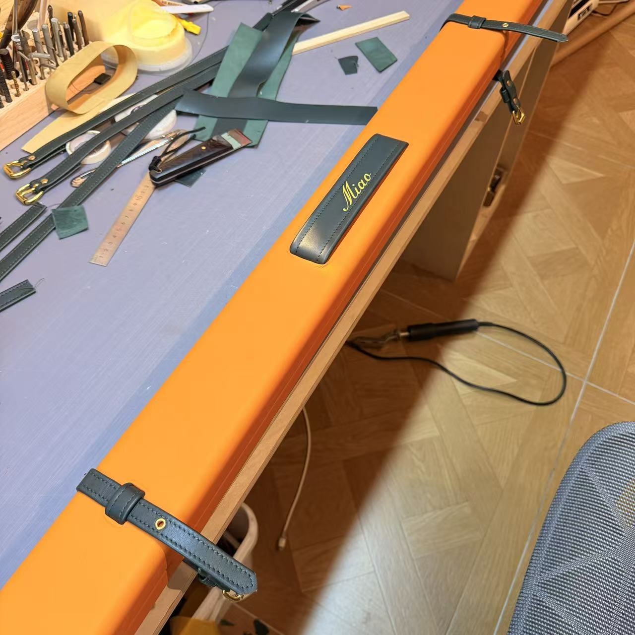Orange leather cue case with green straps and Miao lettering on the workshop bench